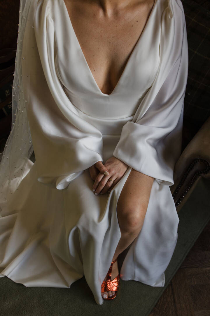 A bride sits in her draped silk wedding dress with long sleeves and a little of her bright orange shoes show under the dress.