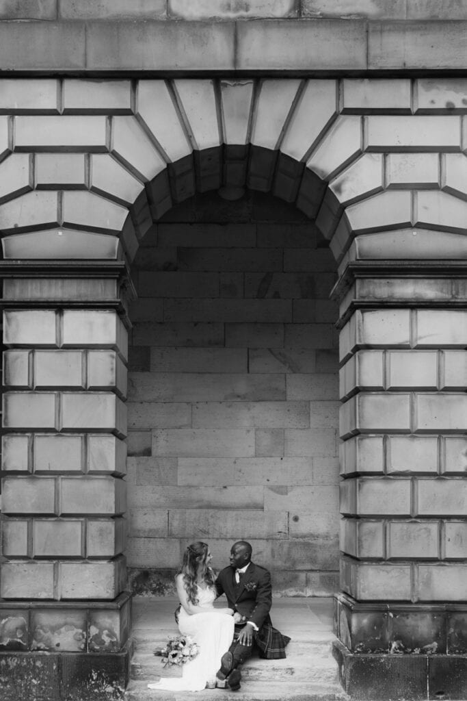 Elegant wedding couple seated beneath historic stone arch at Scottish venue.
