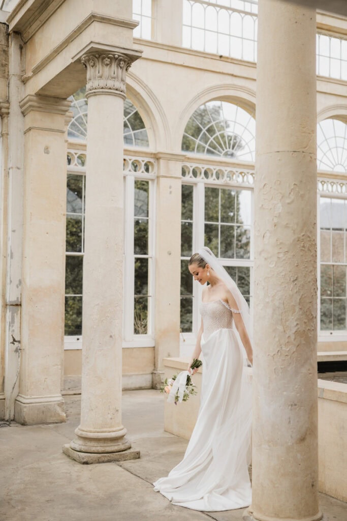 Elegant bride in a white wedding gown inside a historic conservatory with tall columns and large arc.