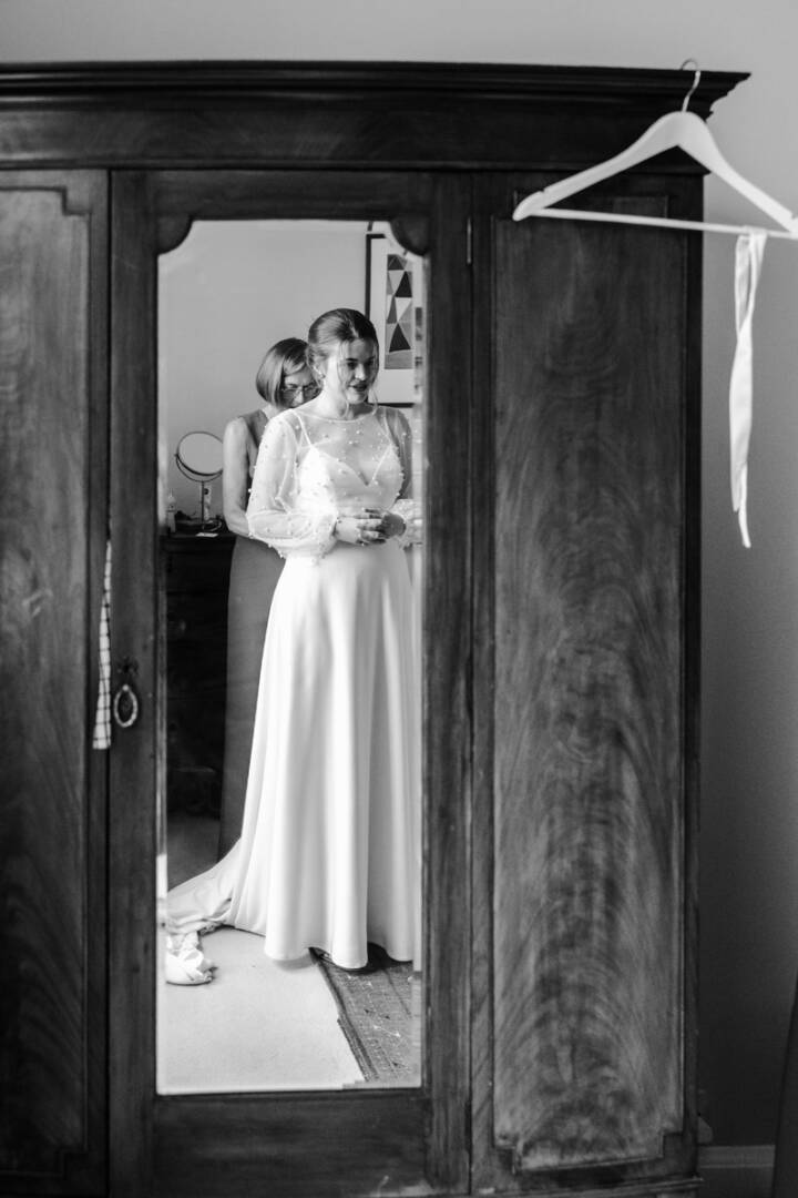 Bride preparing for wedding reflected in vintage wooden wardrobe mirror.