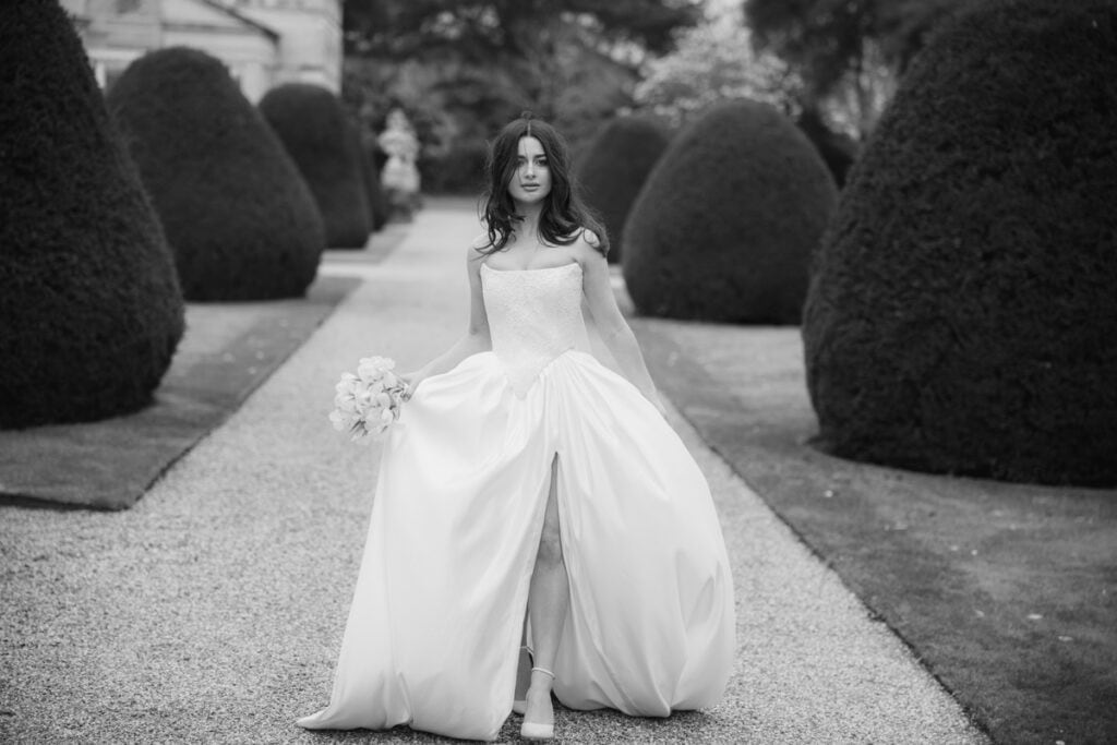 Elegant bride in a wedding gown walking through a formal garden in black and white.