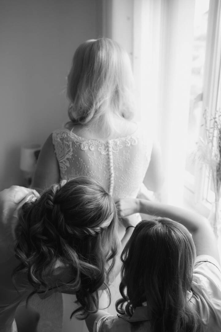 Bride and bridesmaids preparing for wedding by window in black and white.